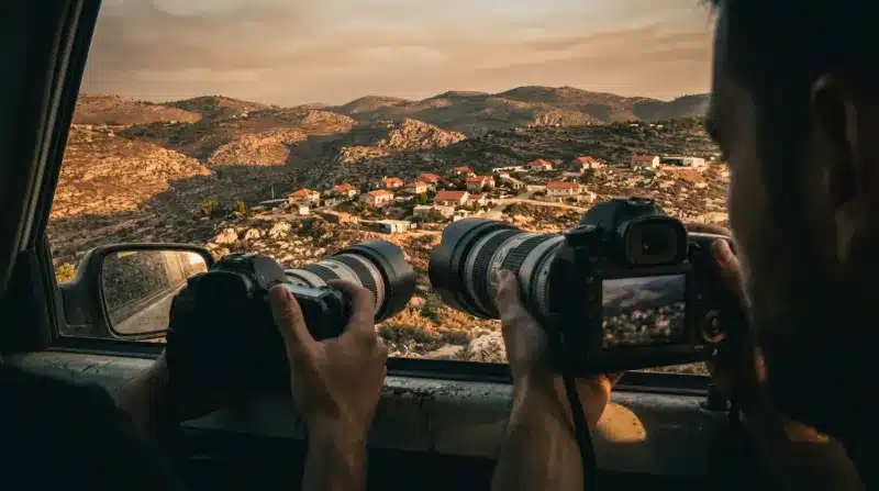 שני צלמים אוחזים במצלמות DSLR עם עדשות טלפוטו ארוכות ומצלמים כפר נופי בשעת השקיעה מתוך חלון של רכב.