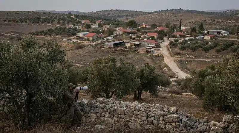 צלם בלבוש צבאי משתמש בעדשת טלפוטו כדי לצלם יישוב בגדה המערבית מעבר לחומת אבן.