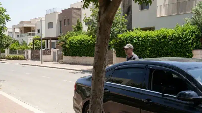 אדם נכנס לרכב שחור ברחוב מגורים עם בתים מודרניים וצמחייה ירוקה