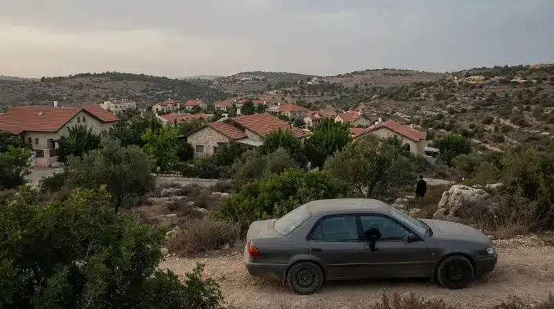 מכונית ישנה חונה בשביל עפר המשקיף על יישוב בעל גגות אדומים בגבעות יהודה ושומרון.