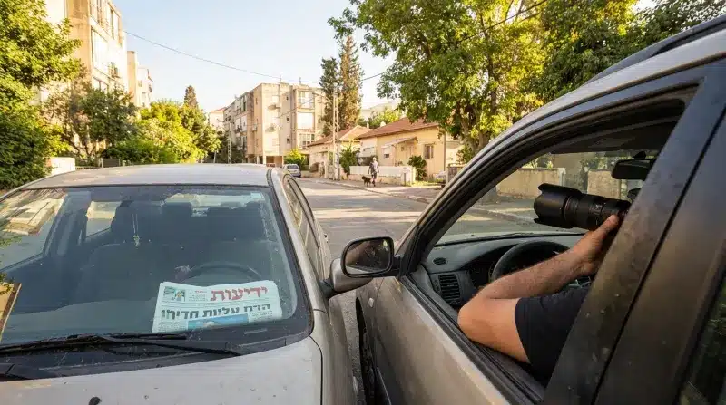 אדם מצלם במצלמה עם עדשת טלפוטו גדולה מתוך חלון של מכונית ברחוב בישראל, כשעיתון "ידיעות אחרונות" מונח על דשבורד של רכב סמוך.