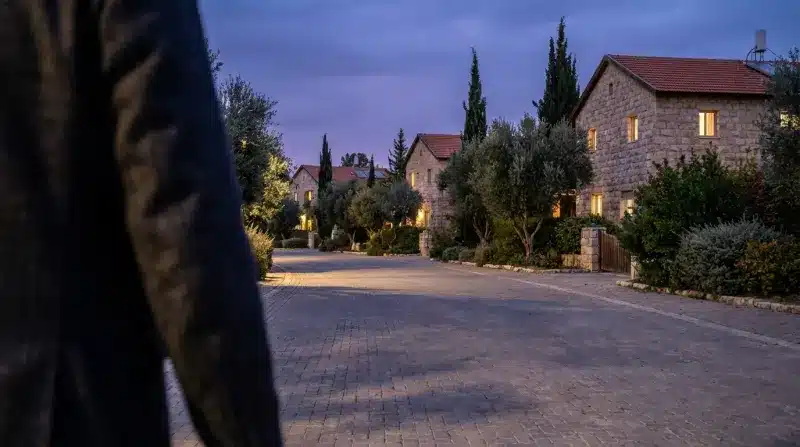 רחוב פסטורלי מרוצף אבן בערב, עם בתי אבן מוארים ועצי זית, מנקודת מבטו של אדם ההולך ברחוב.