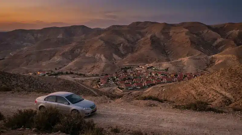מכונית סדאן כסופה חונה בנקודת תצפית על דרך עפר במדבר יהודה, המשקיפה על יישוב מדברי קטן בשעת השקיעה.