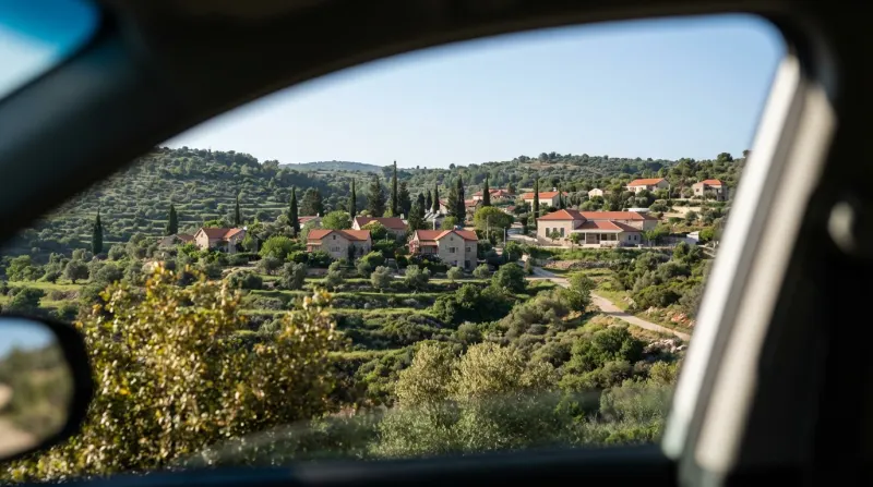 מבט מחלון רכב אל עבר יישוב כפרי פסטורלי עם בתי אבן וגגות רעפים אדומים, השוכן בלב גבעות ירוקות ומדורגות.