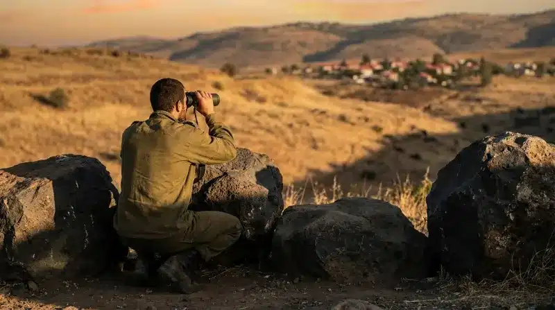 חייל צה"ל כורע מאחורי סלעים ומשקיף במשקפת על יישוב בנוף הררי יבש בשעת שקיעה.