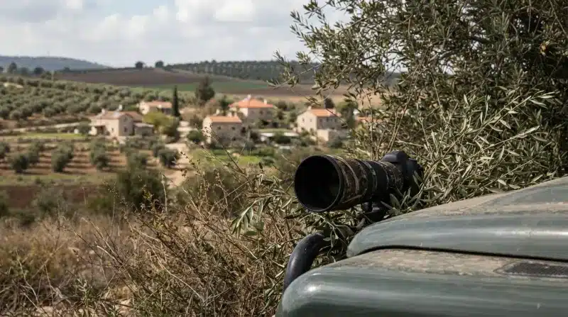 עדשת מצלמה גדולה מוסווית על גבי רכב שטח, משקיפה על נוף כפרי של יישוב ושדות.