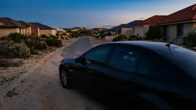 מכונית שחורה חונה על שביל עפר חולי בשכונת מגורים שקטה בקרבת דיונות חול בשעת דמדומים.