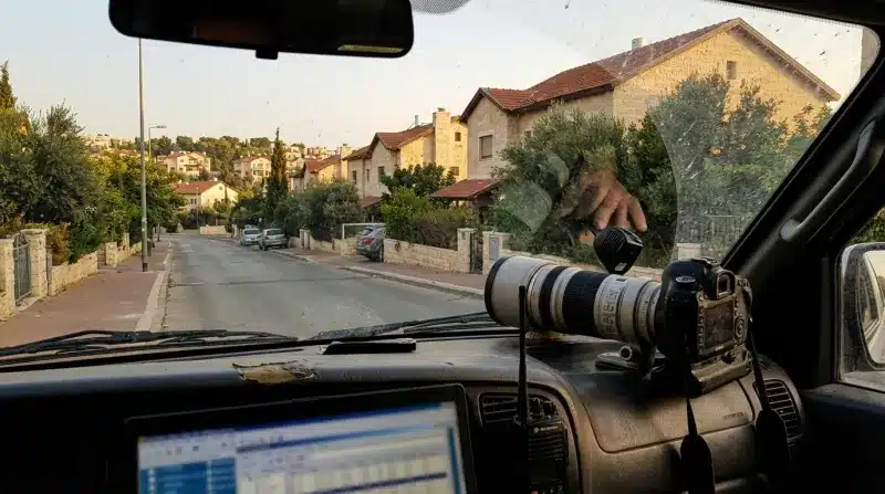 מבט מתוך רכב מעקב: מצלמה מקצועית עם עדשת טלפוטו גדולה, מכשיר קשר ומחשב נייד על הדשבורד ברחוב מגורים.